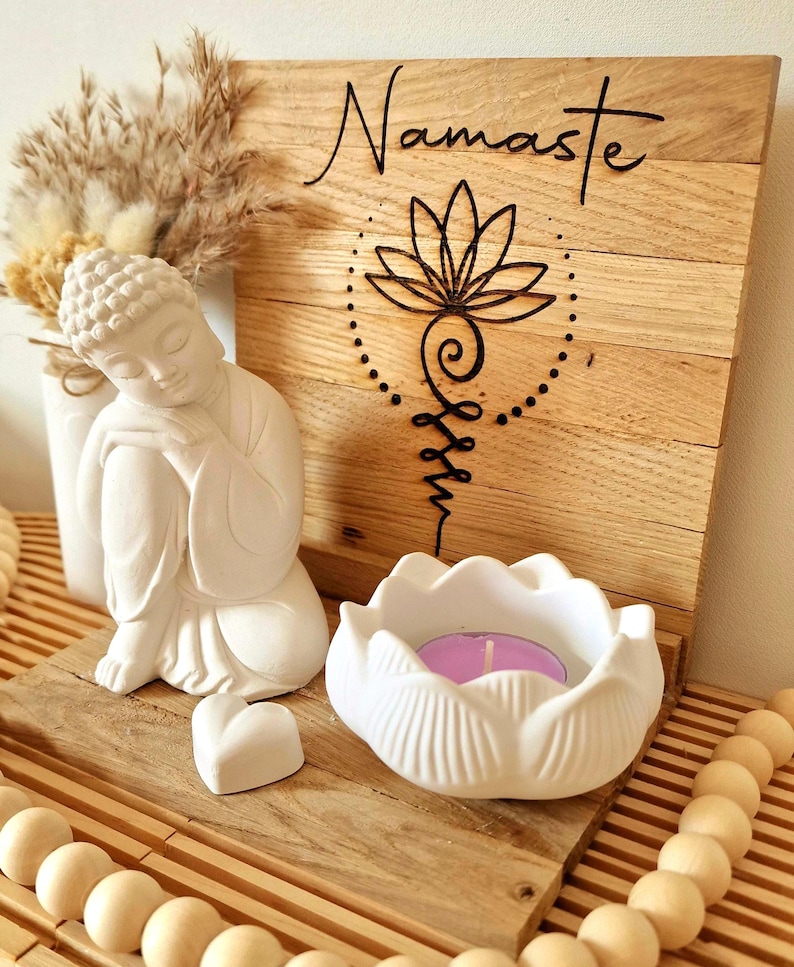May include: A wooden plaque with the word Namaste and a black lotus symbol, featuring a white Buddha figurine, a white lotus-shaped candle holder with a purple tea light, a small white heart, and a dried floral arrangement on a wooden base.