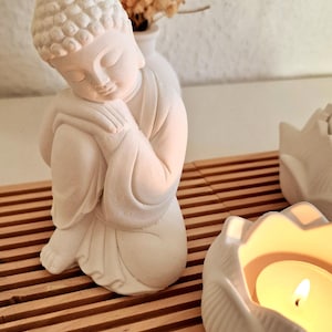 May include: A matte white figurine of a seated Buddha with hands clasped near the face, positioned on a slatted wooden surface next to a white lotus-shaped candle holder containing a lit tea light candle and a small vase with dried flowers.