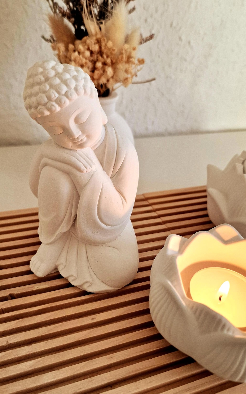 May include: A matte white figurine of a seated Buddha with hands clasped near the face, positioned on a slatted wooden surface next to a white lotus-shaped candle holder containing a lit tea light candle and a small vase with dried flowers.