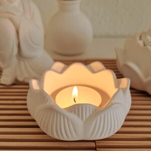 May include: A white ceramic lotus flower candle holder with a matte finish, featuring a lit tea light candle in the centre. The petals have vertical ridges and the holder is shaped like an open blossom, providing a warm glow from the flame.