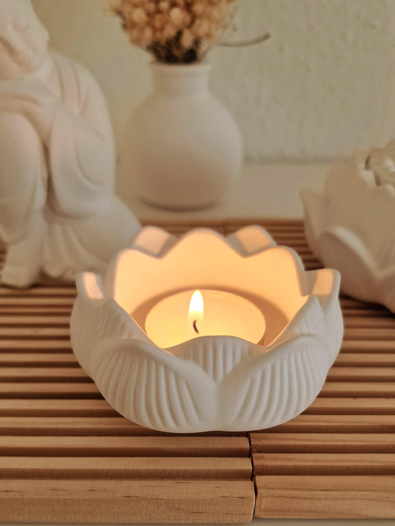 May include: A white ceramic lotus flower candle holder with a matte finish, featuring a lit tea light candle in the centre. The petals have vertical ridges and the holder is shaped like an open blossom, providing a warm glow from the flame.