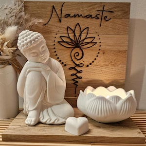 Könnte beinhalten: Eine weiße Buddha-Statue sitzt auf einem Holzregal mit einem herzförmigen Objekt und einem lotusförmigen Kerzenhalter. Im Hintergrund befindet sich ein Holzschild mit dem Wort "Namaste" und einem Lotusblumen-Design.