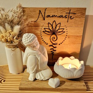 May include: A white Buddha figurine sits beside a white lotus-shaped candle holder with a lit tea light, a small white heart, and a wooden sign featuring the word Namaste and a black lotus symbol, accompanied by a vase of dried beige flowers.