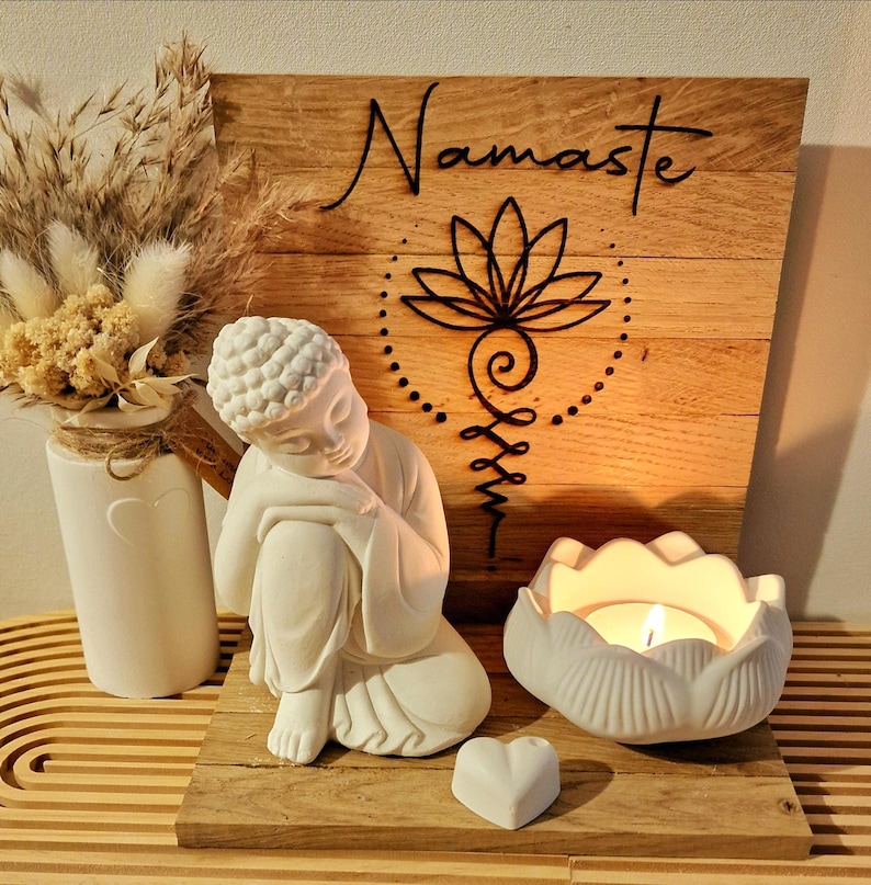 May include: A white Buddha figurine sits beside a white lotus-shaped candle holder with a lit tea light, a small white heart, and a wooden sign featuring the word Namaste and a black lotus symbol, accompanied by a vase of dried beige flowers.