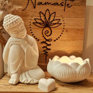 May include: A white Buddha figurine in a seated pose, a white lotus-shaped candle holder with a lit candle, and a small white heart-shaped object. Behind them is a square wooden plaque with the word Namaste and a black lotus line art design.
