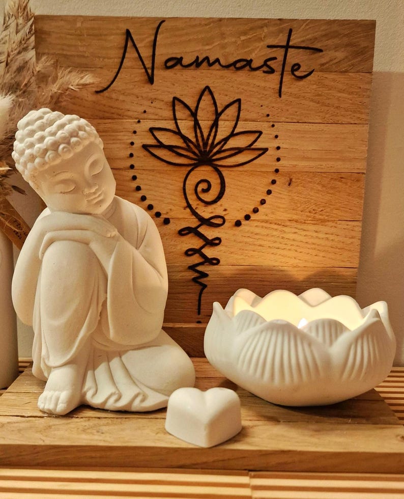 May include: A white Buddha figurine in a seated pose, a white lotus-shaped candle holder with a lit candle, and a small white heart-shaped object. Behind them is a square wooden plaque with the word Namaste and a black lotus line art design.