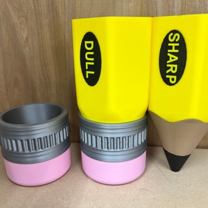 May include: Three pencil-shaped containers. Two are yellow with the words "DULL" and "SHARP" on them. The third is a gray cylinder with a pink base. The pencil-shaped containers are for desk organization.
