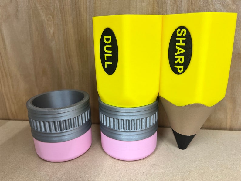May include: Three pencil-shaped containers. Two are yellow with the words "DULL" and "SHARP" on them. The third is a gray cylinder with a pink base. The pencil-shaped containers are for desk organization.