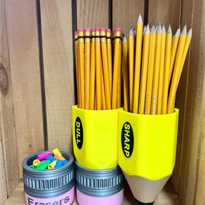 May include: A pencil-themed desk organizer set. Includes a small pink container labeled "Erasers" and two yellow pencil-shaped holders, one labeled "Dull" and the other "Sharp", filled with pencils. The pencil holders are 3D printed.