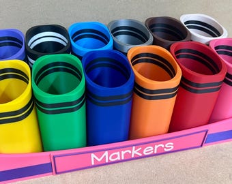 May include: A set of colorful markers in a pink tray. The markers are in various colors, including yellow, green, blue, orange, red, and pink. Each marker has a black stripe around the top. The tray has the word "Markers" on the front.