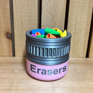 May include: A pink container labeled "Erasers" with a gray rim, filled with colorful eraser caps. The container has a cylindrical shape with a textured design around the middle. The erasers are in various colors, including blue, green, orange, and yellow.