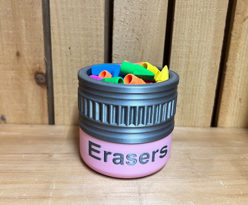 May include: A pink container labeled "Erasers" with a gray rim, filled with colorful eraser caps. The container has a cylindrical shape with a textured design around the middle. The erasers are in various colors, including blue, green, orange, and yellow.