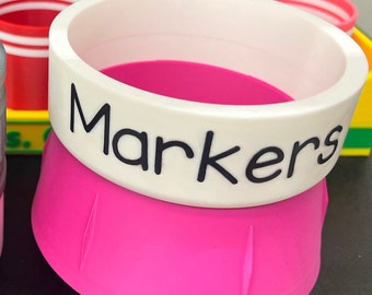 May include: A pink plastic container with a white rim and the word "Markers" written in black. The container is designed to hold markers or other art supplies.