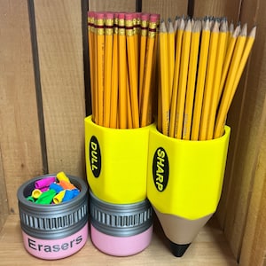 May include: A yellow pencil-shaped desk organizer with the words "DULL" and "SHARP" on the sides, holding numerous yellow pencils. A smaller container labeled "Erasers" holds colorful erasers. The organizer is a fun, back-to-school themed item.