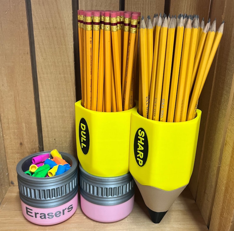 May include: A yellow pencil-shaped desk organizer with the words "DULL" and "SHARP" on the sides, holding numerous yellow pencils. A smaller container labeled "Erasers" holds colorful erasers. The organizer is a fun, back-to-school themed item.