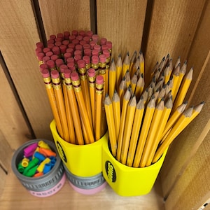May include: A collection of yellow pencils with pink erasers in a yellow pencil-shaped holder. A small container of colorful eraser caps sits nearby. The pencils are arranged in a wooden crate.