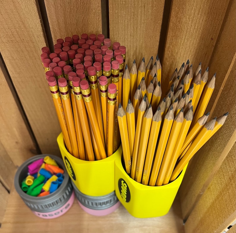 May include: A collection of yellow pencils with pink erasers in a yellow pencil-shaped holder. A small container of colorful eraser caps sits nearby. The pencils are arranged in a wooden crate.