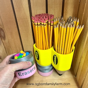 May include: A yellow pencil-shaped desk organizer with the words "Dull" and "Sharp" on the sides, holding numerous yellow pencils with pink erasers. A separate container labeled "Erasers" holds colorful eraser caps. The background is wooden.