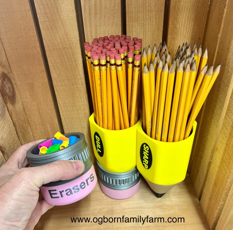 May include: A yellow pencil-shaped desk organizer with the words "Dull" and "Sharp" on the sides, holding numerous yellow pencils with pink erasers. A separate container labeled "Erasers" holds colorful eraser caps. The background is wooden.