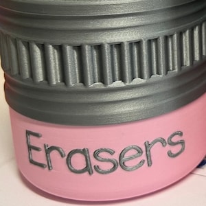 May include: A pink container with the word "Erasers" printed on the side. The top of the container is gray with a ridged design. The container is likely for storing erasers.