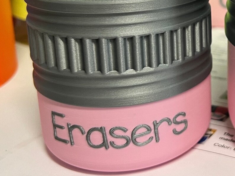 May include: A pink container with the word "Erasers" printed on the side. The top of the container is gray with a ridged design. The container is likely for storing erasers.