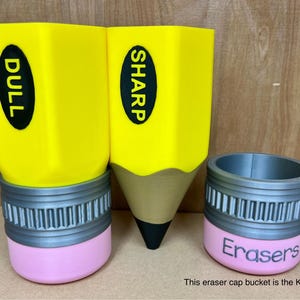 May include: Three pencil-shaped containers. Two are yellow with black text reading "DULL" and "SHARP". The third is a pink and gray eraser cap bucket with the word "Erasers". The pencil containers have a gray band and pink base.