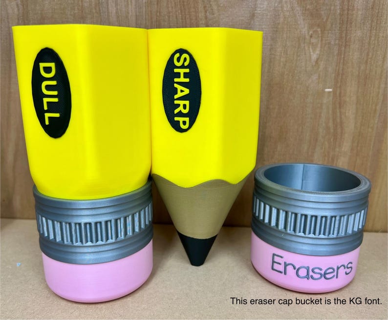 May include: Three pencil-shaped containers. Two are yellow with black text reading "DULL" and "SHARP". The third is a pink and gray eraser cap bucket with the word "Erasers". The pencil containers have a gray band and pink base.
