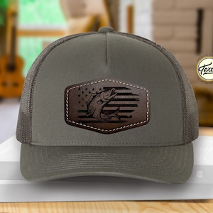 May include: A green and black trucker hat with a brown leather patch featuring a fish jumping over an American flag. The patch has the text "Texas" on it.