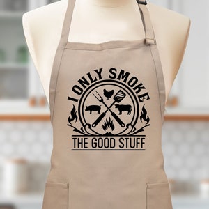 May include: Tan-colored kitchen apron with a black graphic that reads "I Only Smoke The Good Stuff" and features a grill, chicken, steak, and pig.