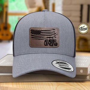 May include: A gray and black trucker hat with a brown leather patch featuring an American flag and a tractor. The patch has a distressed look. The hat has a black mesh back and a gray front. The hat has a Snapback closure.