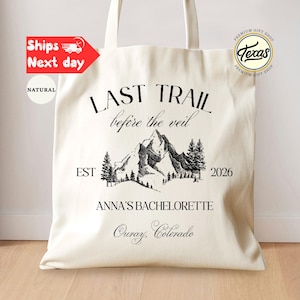 May include: Cream-colored tote bag with black text and a mountain graphic. The text reads "LAST TRAIL before the veil" and "ANNA'S BACHELORETTE Ouray, Colorado 2026". The bag has two handles and is made of a natural fabric.