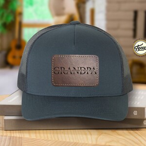 以下が含まれることがあります： ヴィンテージなフォントで「GRANDPA」と書かれた茶色の革のパッチが付いた濃い青色のトラッカーハットです。