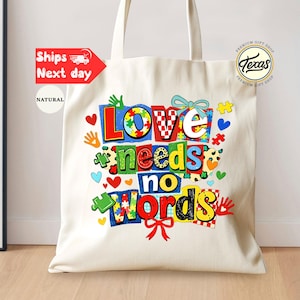 May include: A natural canvas tote bag with the words "Love needs no words" in colorful, block letters. The design includes puzzle pieces, hearts, and handprints. The bag has long handles and is likely for carrying items.