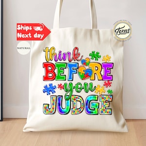 May include: A natural canvas tote bag with the words "think BEFORE you JUDGE" in colorful letters and puzzle piece graphics. The bag has long handles and a simple design, suitable for carrying various items.
