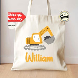 May include: A white canvas tote bag with a yellow excavator design and the name "William" printed in yellow. The bag has a natural fabric texture.