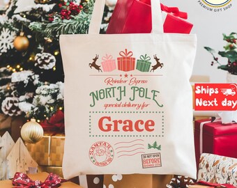 May include: A cream-colored tote bag with a Christmas theme. The bag features the text "Reindeer Express, North Pole, special delivery for Grace." The design includes reindeer, gift boxes, and a Santa Claus stamp. The bag is filled with red gifts.