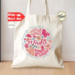 May include: A white canvas tote bag with a floral design and the word "madre" in the center. The floral design is in shades of pink, red, and blue. The text around the floral design reads "Amiga, Incansable, Corprendente, Protectora, Confidente, Amado, Esperanzado".