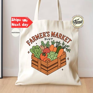 May include: Cream-colored tote bag with the text "FARMER'S MARKET SLUT" in orange and a wooden crate graphic filled with vegetables. The bag has long handles and a "Texas" logo.