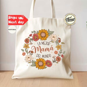 May include: A white canvas tote bag with a floral design and the text 'La Mejor Mamá Del Mundo' in Spanish. The floral design features sunflowers, daisies, and other flowers in shades of yellow, orange, red, and pink. The bag has a natural fabric texture and two handles.