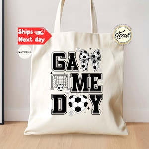 May include: Cream-colored tote bag with the words "GAME DAY" in bold black letters. The design includes soccer balls, a soccer net, and a bow made of soccer balls. A Texas logo is visible.