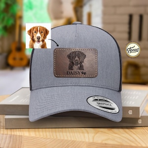 May include: A gray and black trucker hat with a brown leather patch featuring a dog's head and the name "Daisy" on it. The hat has a black mesh back and a snap closure.