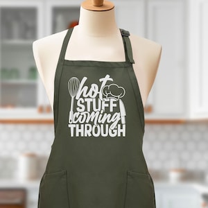 May include: A green apron with two pockets and white text that reads "Hot Stuff Coming Through". The text is accompanied by a chef's hat, a knife, and a whisk.