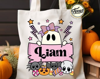 May include: A white canvas tote bag with a pink and purple ghost holding a pink banner with the name "Liam" on it. The ghost has a purple bow and is surrounded by Halloween-themed graphics, including candy corn, a pumpkin, a spider, and a skull.