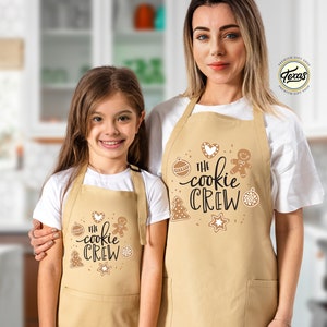 May include: Two people wearing tan aprons with the text "The Cookie Crew" and gingerbread cookie illustrations. The aprons have a pocket on the bottom.