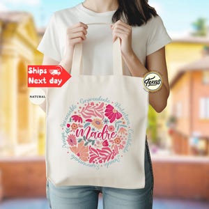 May include: A white canvas tote bag with a floral design and the word "madre" in the center. The design includes pink, red, and blue flowers and leaves. The words "Invaluable,  Incomparable,  Amazing,  Comprehends,  Protective,  Constant,  Unique,  Essential" are written in a circle around the word "madre".