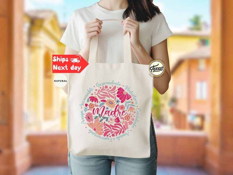 May include: A white canvas tote bag with a floral design and the word "madre" in the center. The design includes pink, red, and blue flowers and leaves. The words "Invaluable,  Incomparable,  Amazing,  Comprehends,  Protective,  Constant,  Unique,  Essential" are written in a circle around the word "madre".