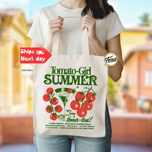 Peut inclure: Un sac fourre-tout en toile naturelle avec le texte "Tomato-Girl SUMMER" en vert. Le design comprend un verre &agrave; martini avec des tomates cerises et la phrase "let's make a toma-tini !" Les d&eacute;tails de la recette sont imprim&eacute;s en dessous.