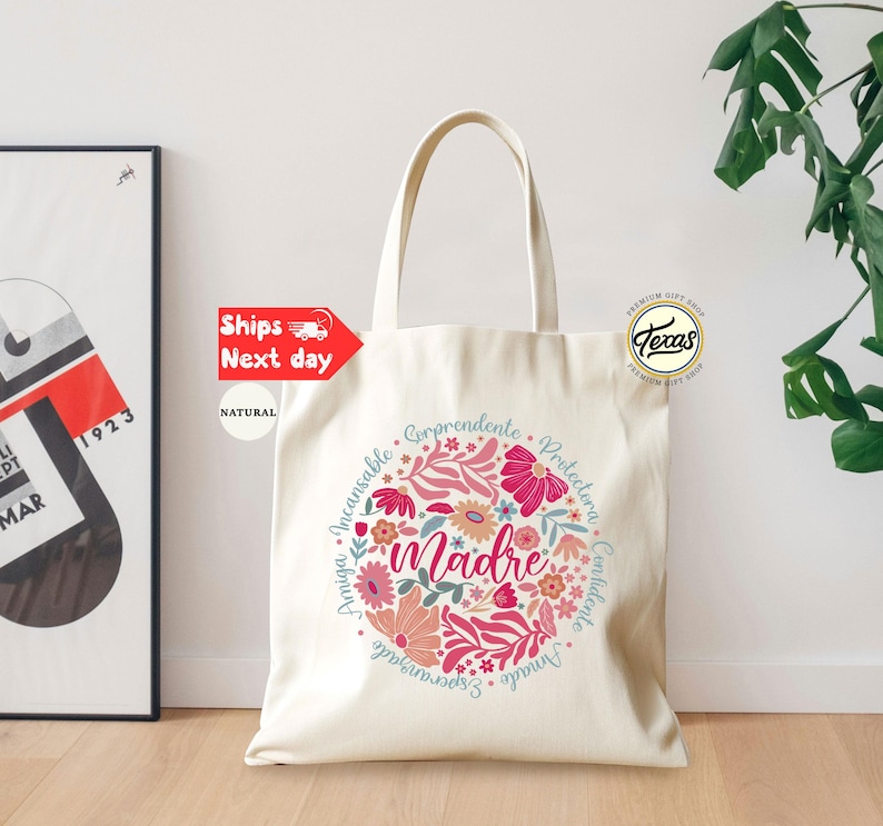 May include: A white canvas tote bag with a floral design and the word "madre" in the center. The floral design is in shades of pink, red, and blue. The text around the floral design reads "Amiga, Incansable, Corprendente, Protectora, Confidente, Amado, Esperanzado".