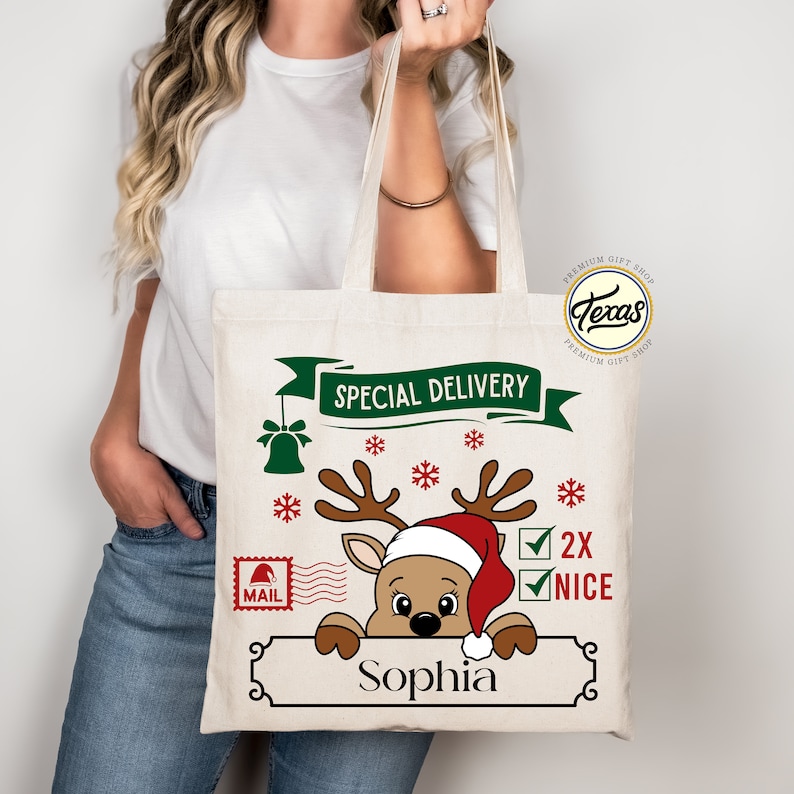 May include: A beige canvas tote bag featuring a reindeer wearing a Santa hat, with the text "Special Delivery." The bag includes the name "Sophia" and the words "2X NICE" with checkmarks, along with snowflakes and a bell.