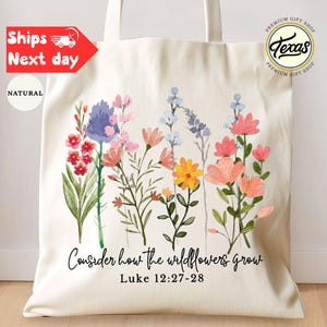 May include: A natural canvas tote bag featuring a watercolor floral design in shades of pink, purple, yellow, and blue. The text "Consider how the wildflowers grow" and Bible verse Luke 12:27-28 are printed below the flowers. The bag has two handles.